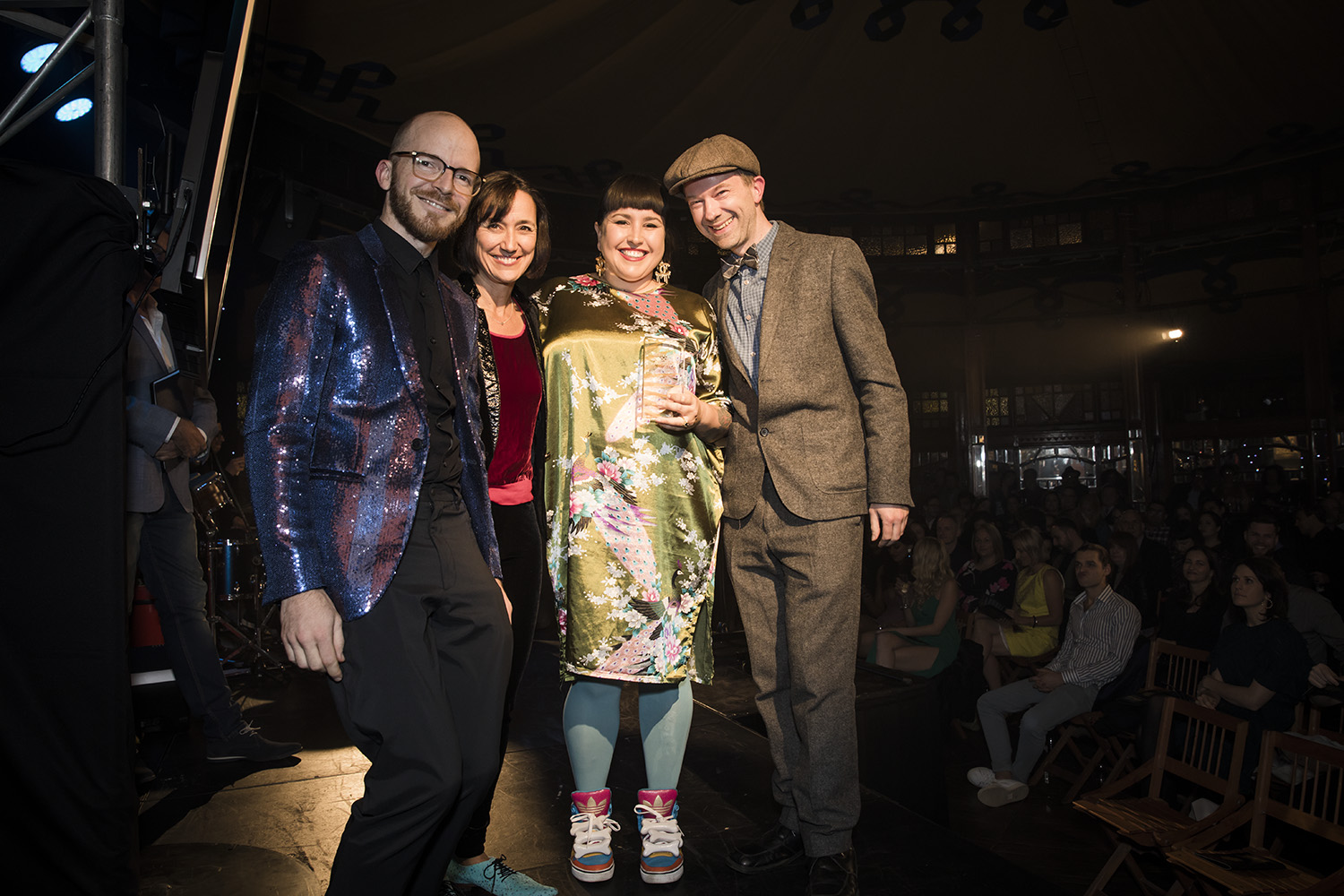 Phil, Carla and Ron accepting the Arts and Entertainment award at the 2015 Australia Podcast Awards from Claudia Taranto. All four are standing and smiling to camera, Carla is holding the award.