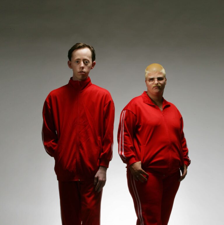 Two performers stand facing the camera, both are in red Adidas tracksuits with white stripes. They stand in front of a gray background.