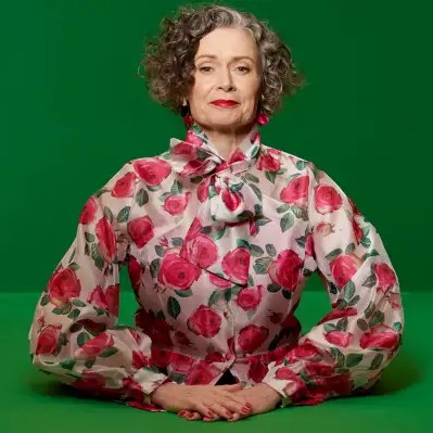 Judith Lucy in a floral shirt is facing the camera smiling in front of a green background.