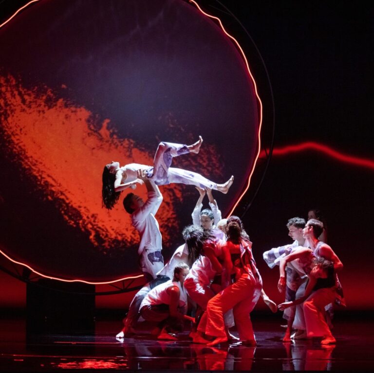 Image from The Hum. A group of dancers perform in front of a large round screen. The stage is black and the round screen is lit with red light. They are raising a dancer on their back, in the middle of a circle.