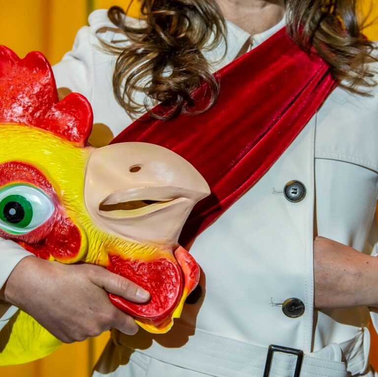 Image is closely cropped on a woman's chest in a military style overcoat, red sash and hand in the breast of the jacket. She is holding a rubber chicken mask.