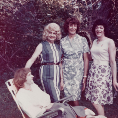 A photo from the 60's with 4 trans women dressed in casual sun dresses,. Their wigs are beautifully styled and they are smiling at the camera. One of the women is on a an outdoor chair smiling back at them.