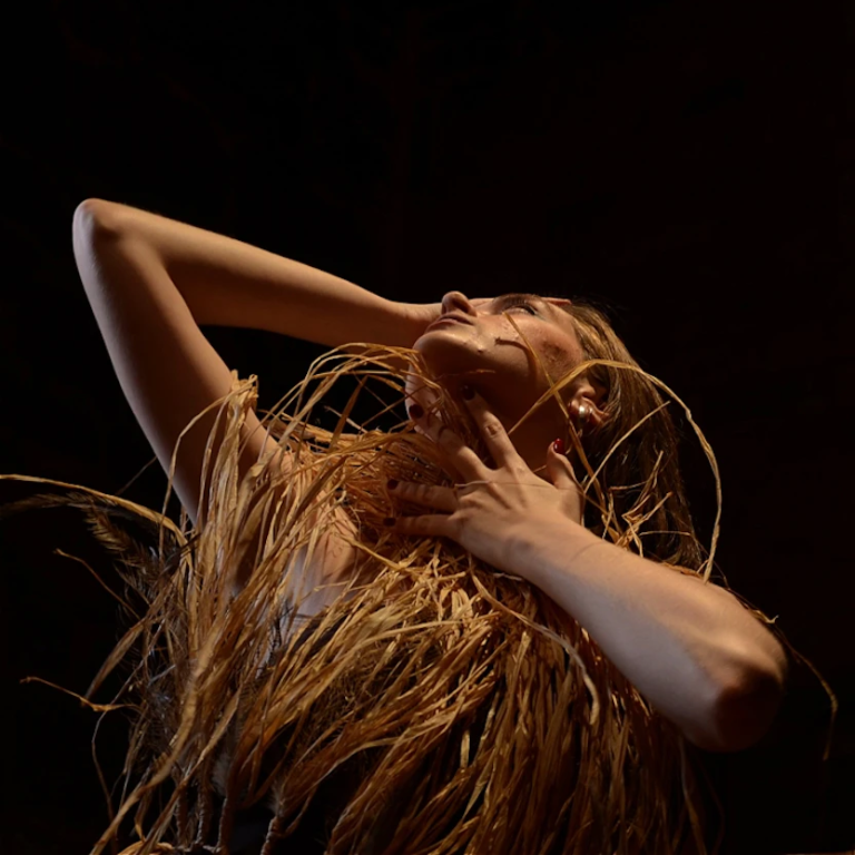 A close up image of Amelia's face and upper body. She is holding grass-straw and emu feathers up to her neck, with her head back and with her hand on her face as she looks up into the dark space around her and the light shines on her.